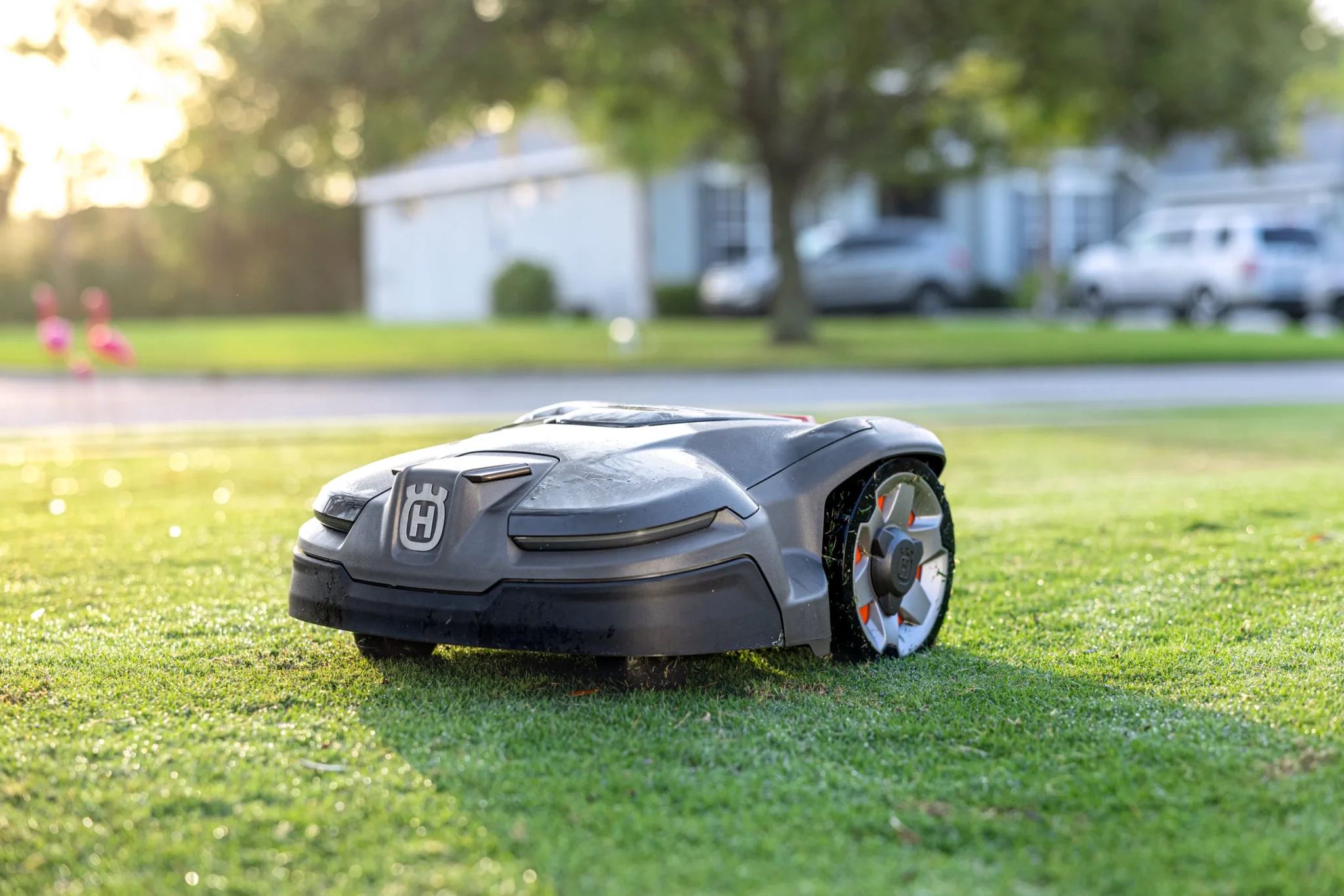 Trädgårdmaskiner i toppklass En robotgräsklippare med Husqvarnas logotyp rör sig över en gräsmatta med hus, träd och parkerade bilar suddiga i bakgrunden.