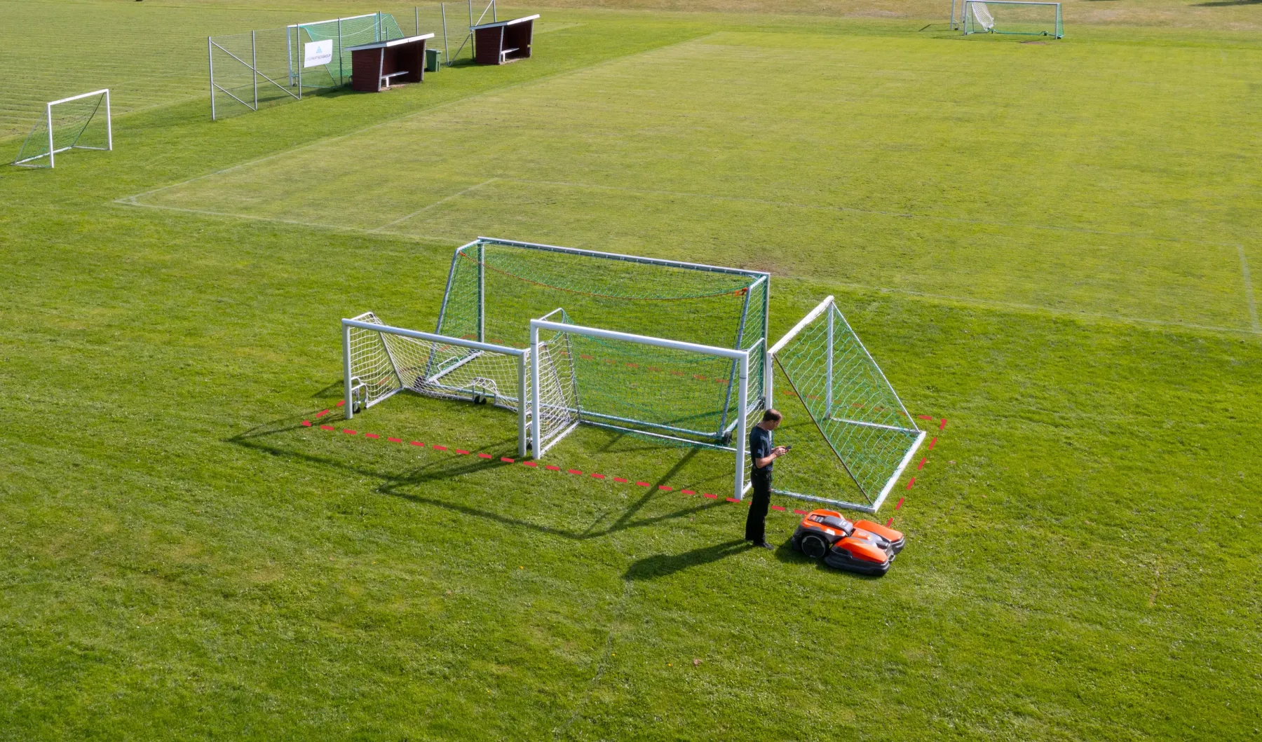 En person använder en robotgräsklippare för att klippa gräs bredvid flera staplade fotbollsmål på en stor, välskött idrottsplats.