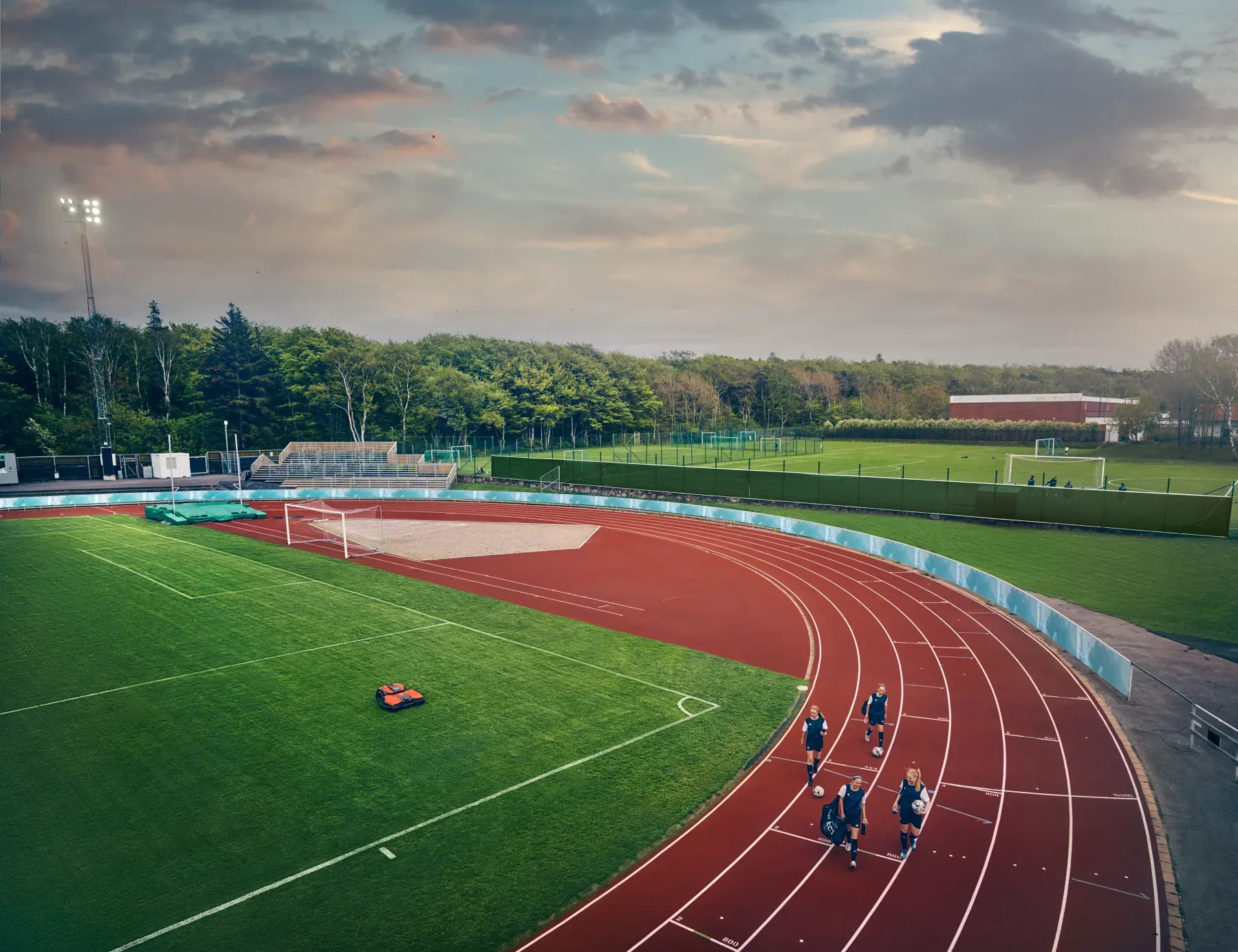 En grupp människor joggar på en röd löparbana på en utomhusstadion bredvid gräsbeklädda sportfält, omgivna av träd under en delvis molnig kvällshimmel.