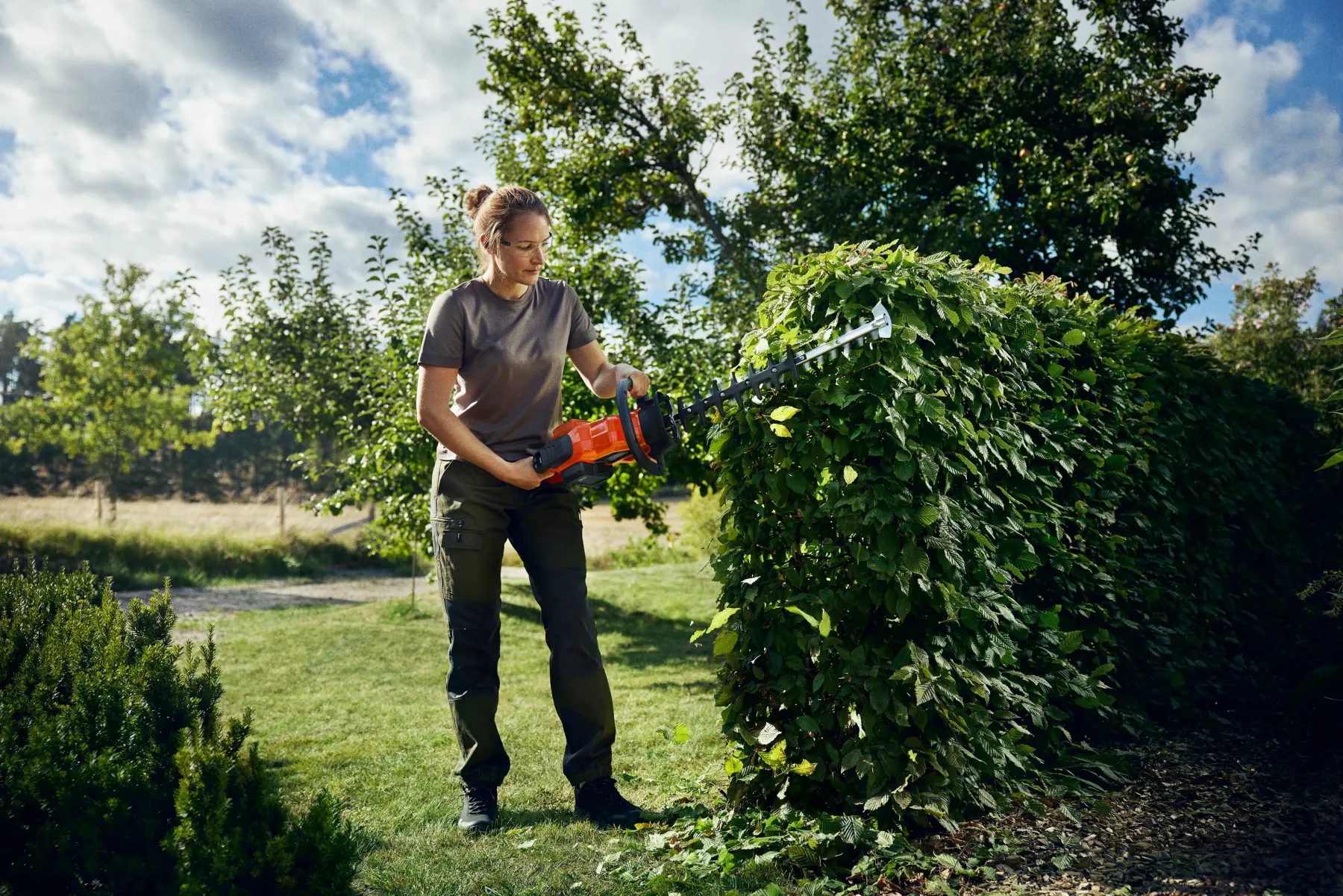 En person använder en orange häcksax för att klippa en hög, lövgrön häck i en trädgård med träd och gräs under en delvis molnig himmel.
