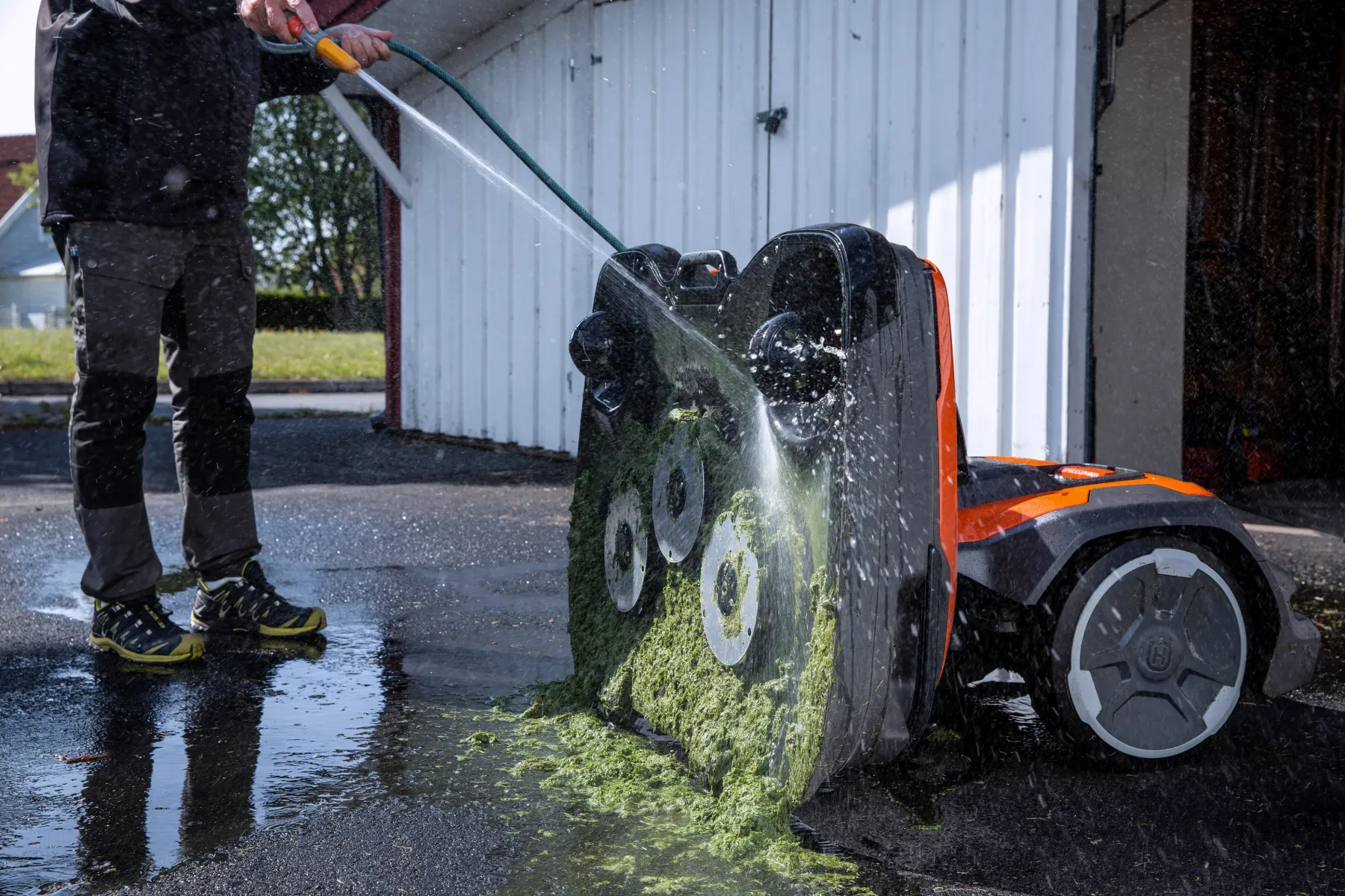 En person använder en slang för att tvätta bort gräsklipp från undersidan av en orange robotgräsklippare på en uppfart utanför ett vitt garage.