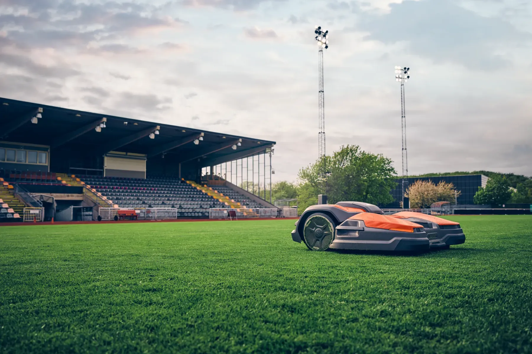 Två orange och grå robotgräsklippare kör på en grön gräsplan i en tom stadion med täckta sittplatser och höga strålkastare, under en molnig himmel.