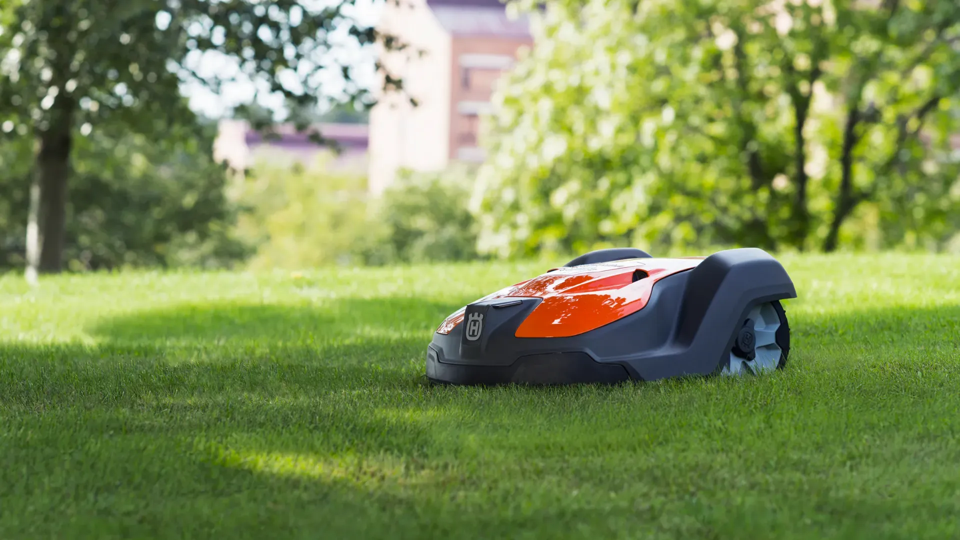Robotgräsklippare för fastighetsbolag En röd och svart robotgräsklippare rör sig över en gräsmatta med gröna träd och suddiga byggnader i bakgrunden.