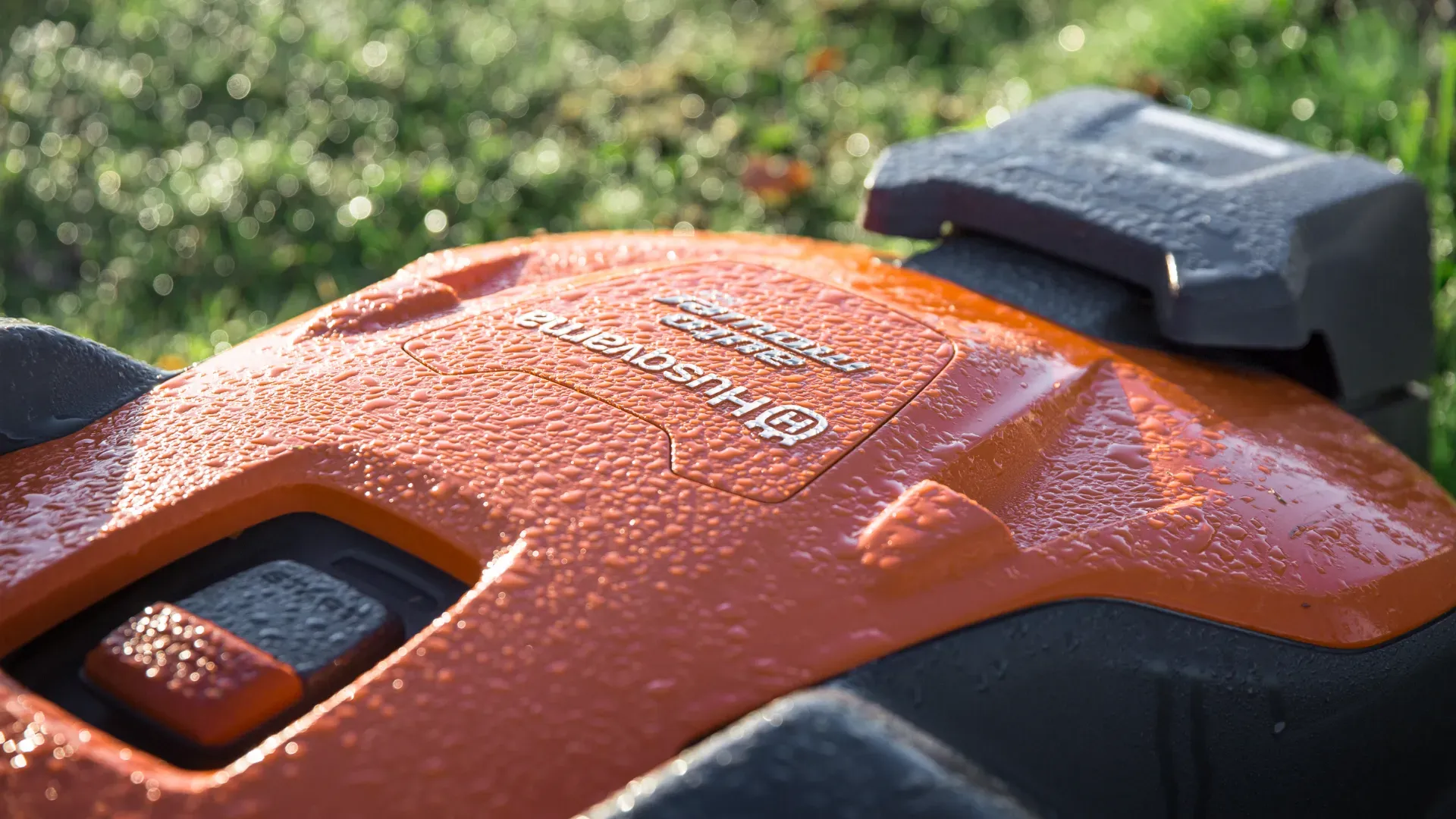imgi_27_robotdetaljbild Närbild av en daggtäckt orange Husqvarna robotgräsklippare med gräs suddigt i bakgrunden. Husqvarnas logotyp och modellinformation syns på maskinens yta.
