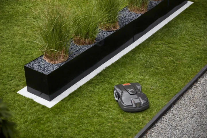Automower-305-uai-720x480 En robotgräsklippare trimmar gräset längs kanten av en modern trädgård med en upphöjd svart planteringskärl fylld med höga gröna växter och mörkt grus.