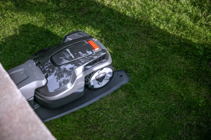 Automower-315-mark-2-uai-720x480 En robotgräsklippare är placerad på grönt gräs intill en sten- eller betongkant, delvis i skuggan, och förbereder sig för att klippa gräsmattan.