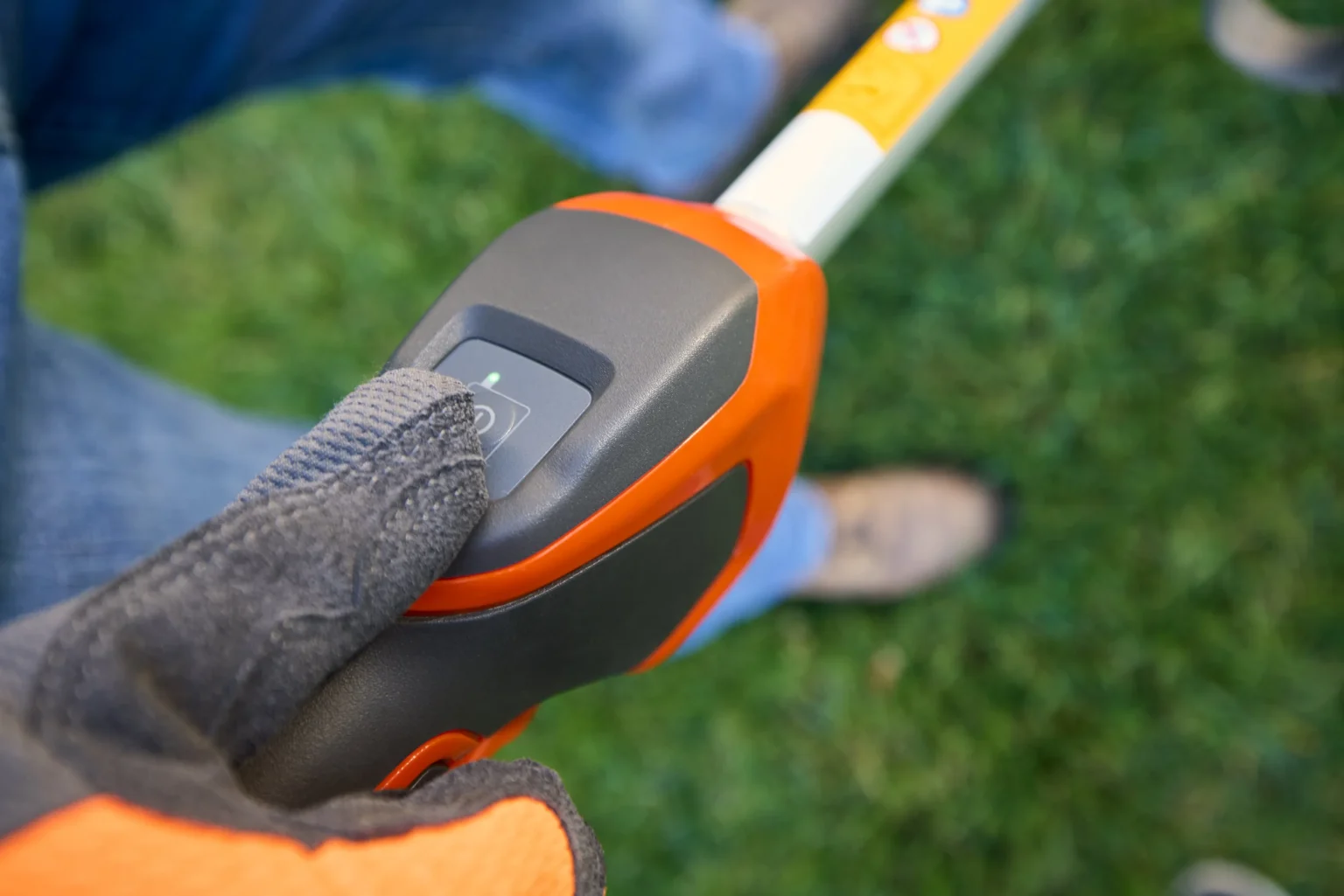 En handskbeklädd hand trycker på strömbrytaren på handtaget till ett orange och svart elverktyg, med grönt gräs och en persons fot synlig i den suddiga bakgrunden.
