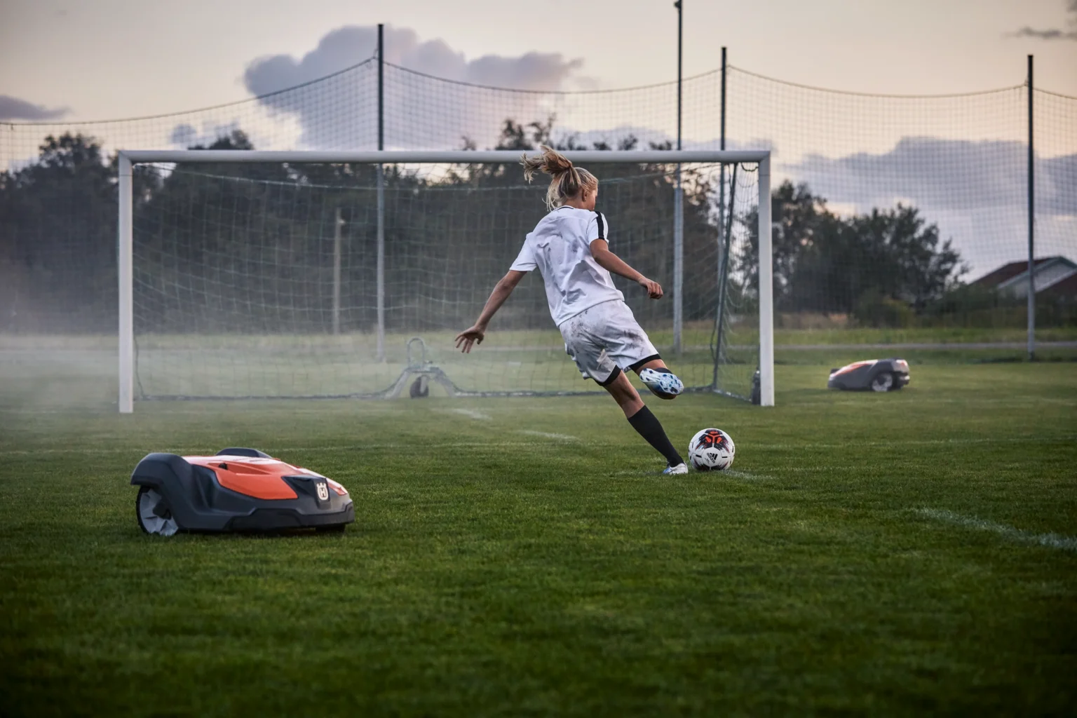 En ung fotbollsspelare i vit uniform sparkar en boll mot ett mål på en gräsplan, medan två robotgräsklippare lugnt sköter om gräset i närheten.