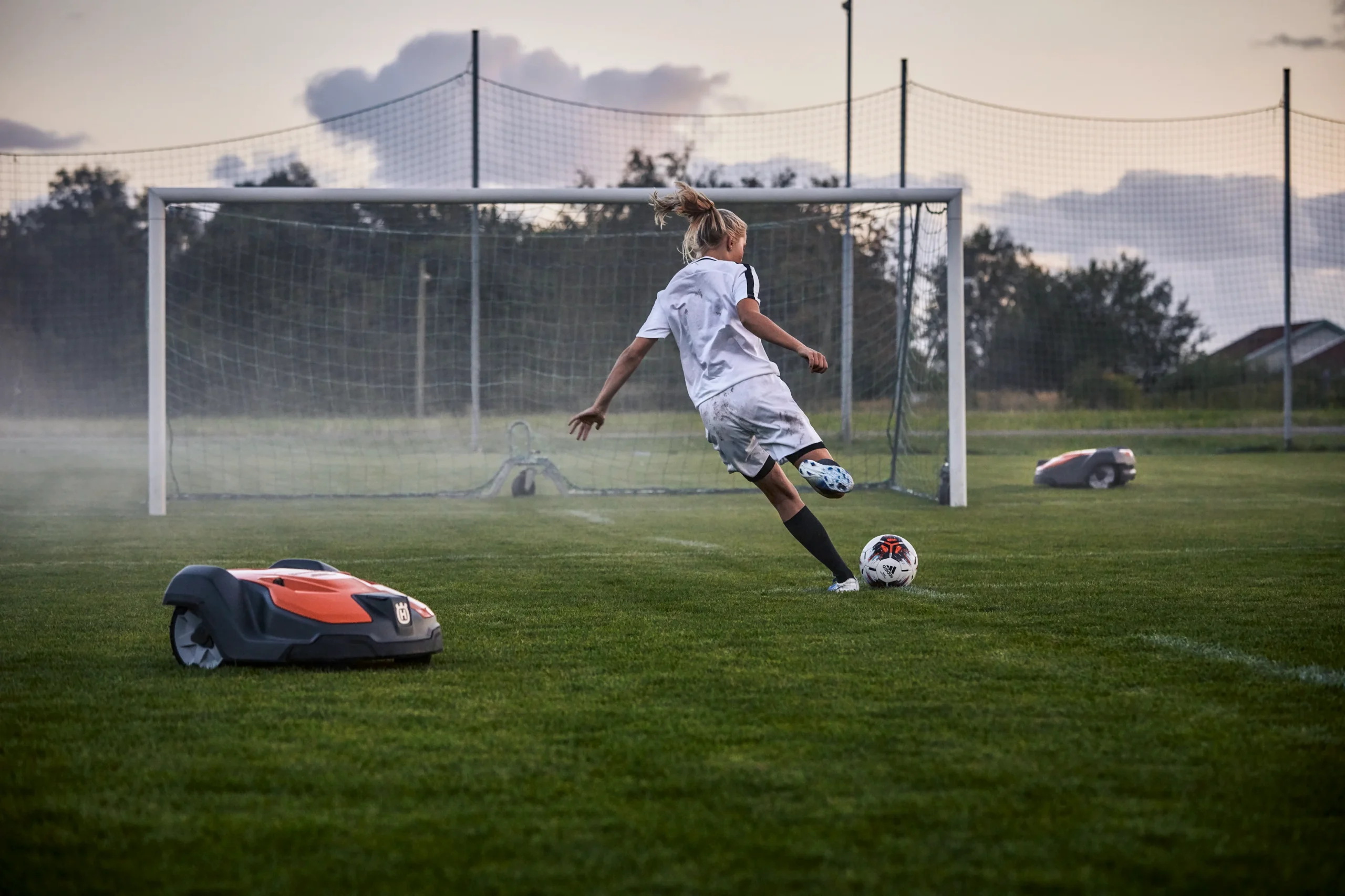 En ung fotbollsspelare i vit uniform sparkar en boll mot ett mål på en gräsplan, medan två robotgräsklippare lugnt sköter om gräset i närheten.