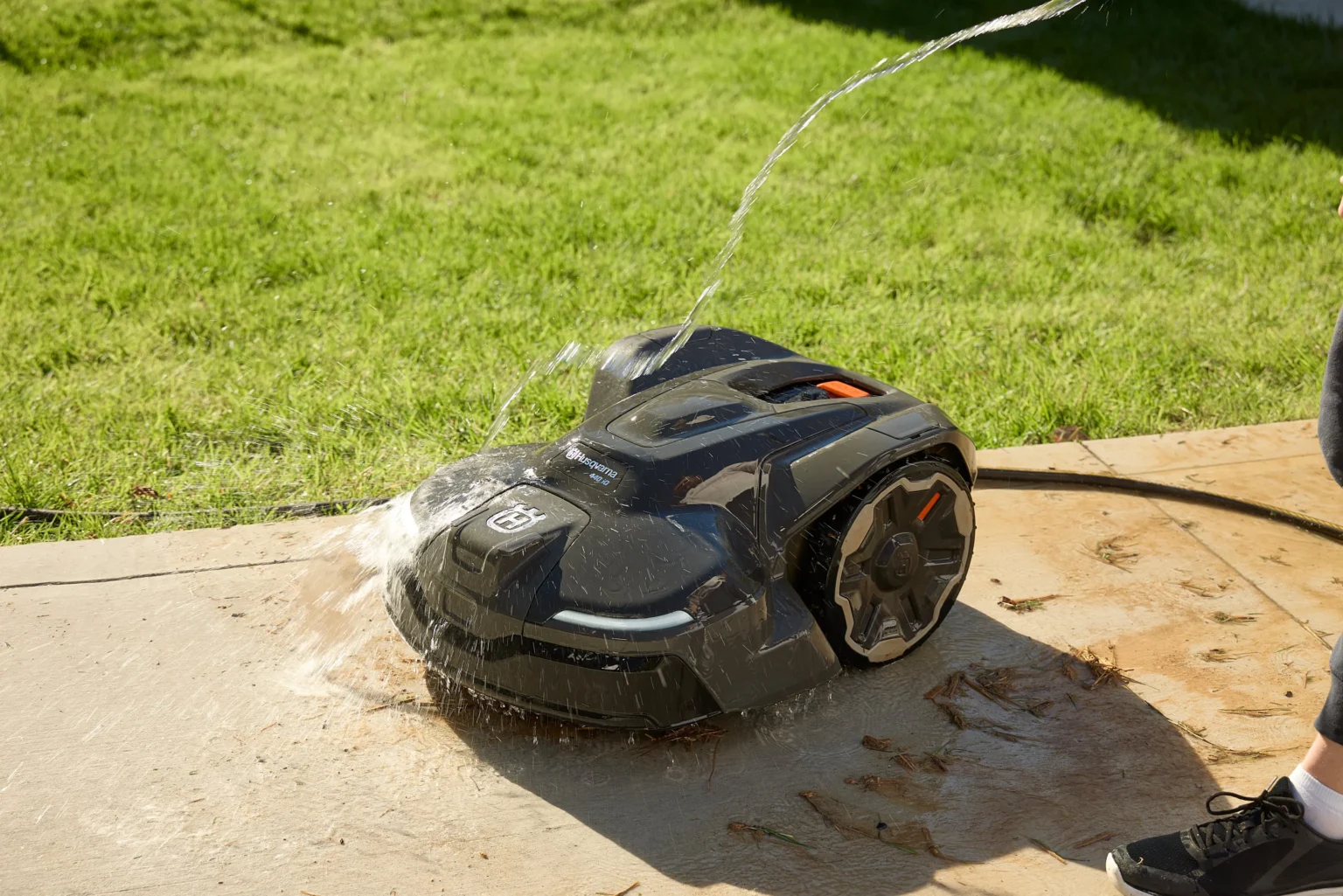 En robotgräsklippare sprutas med vatten från en slang på en betongyta bredvid en gräsmatta, vilket visar dess vattentäta kapacitet.