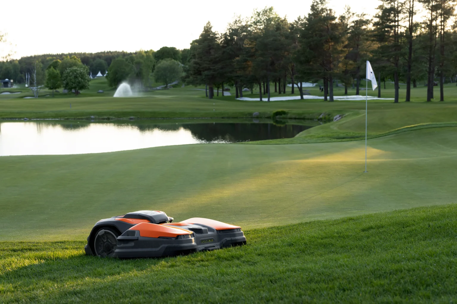 Två orange och svarta robotgräsklippare står på gräset nära en golfgreen med en flagga. En damm, träd, en fontän och avlägsna golfhål syns i bakgrunden i kvällsljus.