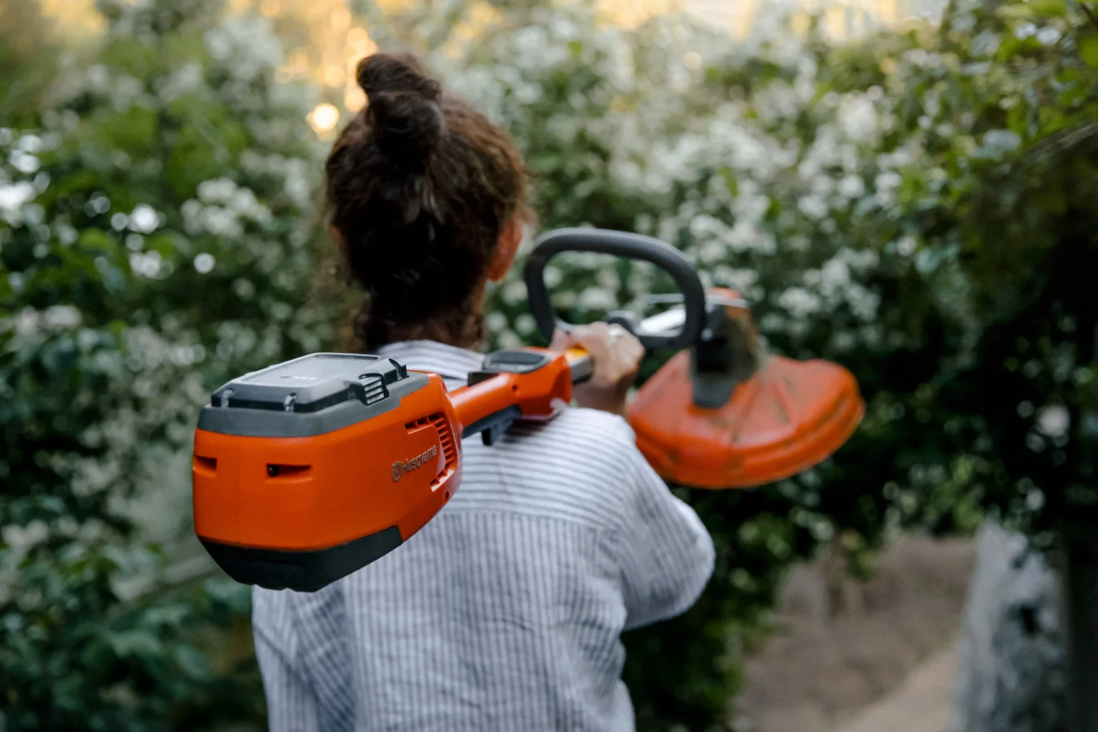 En person med lockigt hår i en knut, klädd i en randig skjorta, bär en orange snortrimmer över axeln medan han går genom en trädgård med gröna buskar och vita blommor.