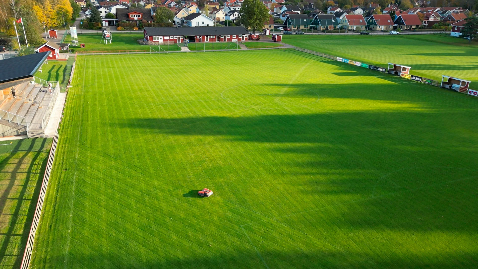 Flygfoto över en grön, välskött fotbollsplan med tomma läktare till vänster och en liten röd robotgräsklippare i mitten; hus och träd omger planen i bakgrunden.