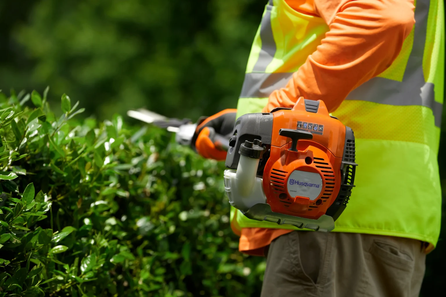 En person i orange skjorta och gul säkerhetsväst trimmar gröna buskar med en orange och svart Husqvarna häcksax i en utomhusmiljö, perfekt för dem som vill köpa en häcksax för sina egna trädgårdsuppgifter.