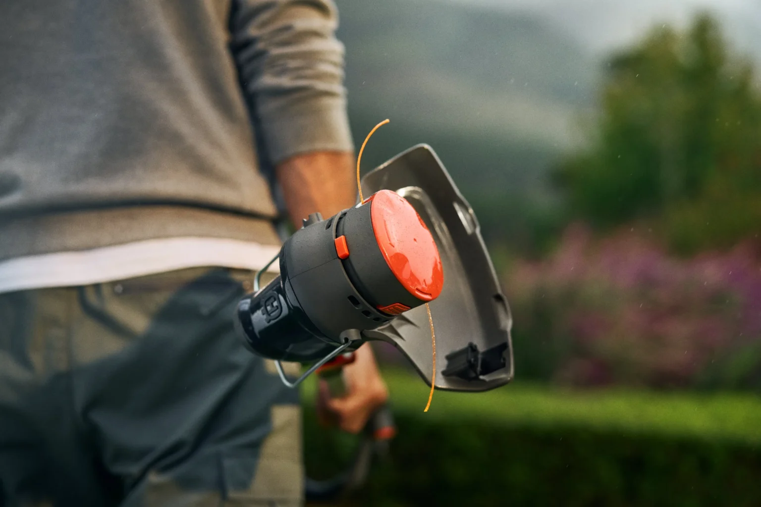 En person håller i en trimmer i en trädgård, med fokus på verktygets orange och svarta skärhuvud och suddig grönska i bakgrunden.
